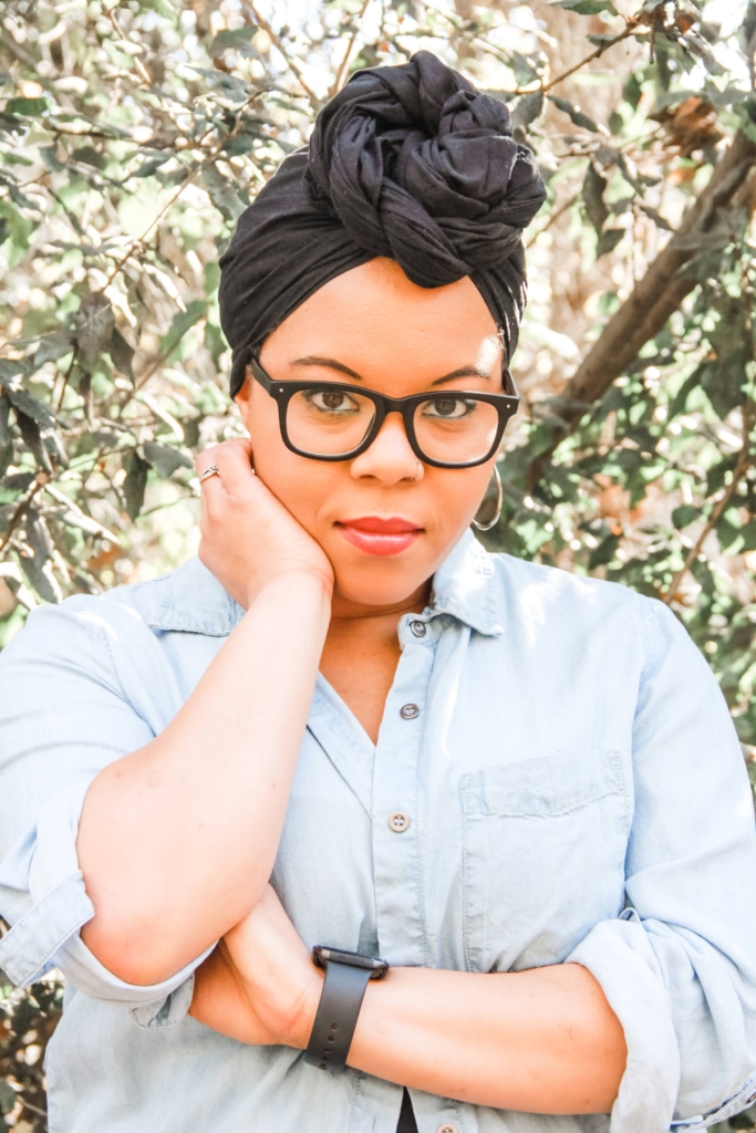 Ashlea, Black Light-Skinned Woman looks directly at the camera. They are wearing a light blue denim shirt, buttoned down in the front. They are wearing a black headwrap with a knot tied to the front and thick black-rimmed glasses.