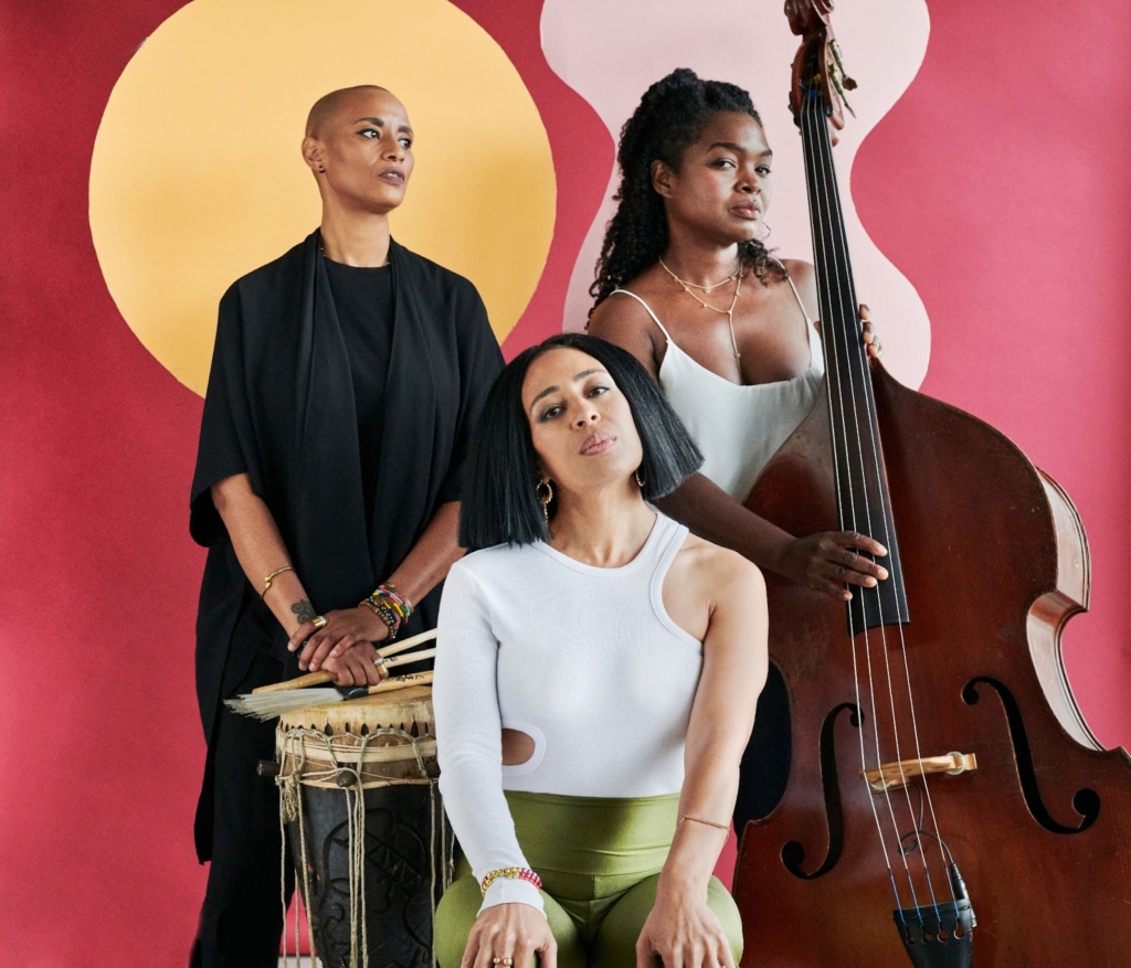 Three Black women musicians, two standing, and one sitting in front, pose for the camera. The two standing have instruments in their hands. 